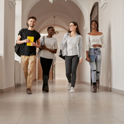Full length portrait of four mixed-race group of young smiling people,casual dressed, walking through the bright university corridor. Happy attractive students are going home after finishing lessons.